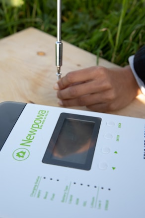 A person's hand is seen using a tool on a wooden surface adjacent to a device labeled 'Newpowa' with buttons and a screen. The background features green grass, indicating an outdoor setting.