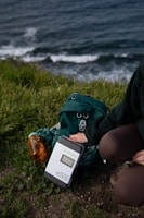 A person kneels on the grass near a coastal area, setting up a solar charge controller labeled 'Newpowa'. Next to the device is a green hiking backpack and a transparent orange water bottle. The background displays ocean waves crashing against the shore.