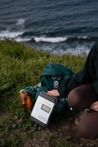 A person kneels on the grass near a coastal area, setting up a solar charge controller labeled 'Newpowa'. Next to the device is a green hiking backpack and a transparent orange water bottle. The background displays ocean waves crashing against the shore.