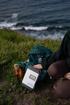 A person kneels on the grass near a coastal area, setting up a solar charge controller labeled 'Newpowa'. Next to the device is a green hiking backpack and a transparent orange water bottle. The background displays ocean waves crashing against the shore.