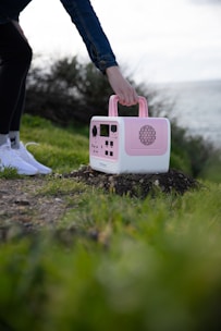 A person is holding a pink and white portable device with multiple dials and speakers, placed on a grassy and rocky outdoor ground near a body of water. The person is wearing white shoes and a blue jacket.