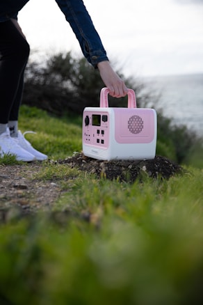 A person is holding a pink and white portable device with multiple dials and speakers, placed on a grassy and rocky outdoor ground near a body of water. The person is wearing white shoes and a blue jacket.