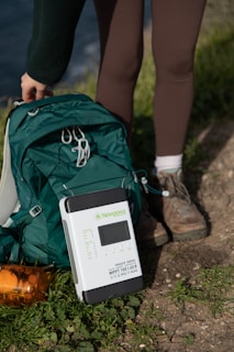 A portable solar charge controller placed on the ground next to a green backpack and an orange water bottle. A person wearing brown leggings and hiking boots is standing nearby, suggesting a hiking or outdoor activity setting.