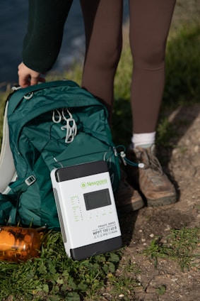 A portable solar charge controller placed on the ground next to a green backpack and an orange water bottle. A person wearing brown leggings and hiking boots is standing nearby, suggesting a hiking or outdoor activity setting.
