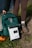 A portable solar charge controller placed on the ground next to a green backpack and an orange water bottle. A person wearing brown leggings and hiking boots is standing nearby, suggesting a hiking or outdoor activity setting.