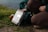 A person is kneeling on the ground next to a green backpack. Beside the backpack, there is an electronic device with a screen and buttons labeled 'Newdome' and 'PRIVATE SERIES MPPT 100 | 40 B'. An orange bottle is partially visible lying on the grass, which leads to the edge of a water body.