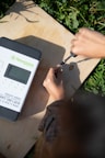 A person is using a screwdriver to adjust something on a wooden surface. Next to them is a solar charge controller labeled 'Newpowa' with details about its power specifications. The scene is bright, with natural light illuminating the area and surrounding greenery visible.