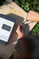 A person is using a screwdriver to adjust something on a wooden surface. Next to them is a solar charge controller labeled 'Newpowa' with details about its power specifications. The scene is bright, with natural light illuminating the area and surrounding greenery visible.