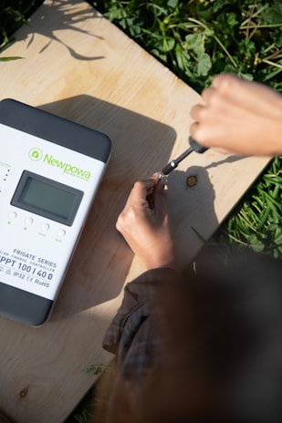A person is using a screwdriver to adjust something on a wooden surface. Next to them is a solar charge controller labeled 'Newpowa' with details about its power specifications. The scene is bright, with natural light illuminating the area and surrounding greenery visible.