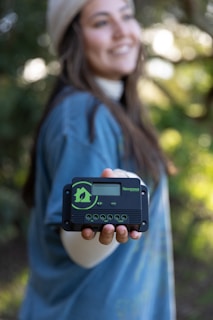 A person with long hair wearing a beanie and a blue shirt holds out a small, rectangular black device with a digital display screen and buttons. The background is blurred, featuring greenery and sunlight, creating a serene and natural setting.