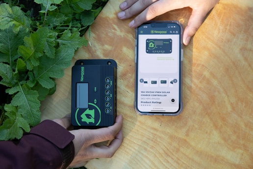 Two hands are holding objects next to some green plants. One hand holds a black and green electronic device with the brand 'Newpowa' displayed. The other hand holds a smartphone showing a product page for a 'Newpowa' 12V/24V PWM Solar Charge Controller, including product ratings and details.