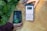 Two hands are holding objects next to some green plants. One hand holds a black and green electronic device with the brand 'Newpowa' displayed. The other hand holds a smartphone showing a product page for a 'Newpowa' 12V/24V PWM Solar Charge Controller, including product ratings and details.