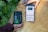 Two hands are holding objects next to some green plants. One hand holds a black and green electronic device with the brand 'Newpowa' displayed. The other hand holds a smartphone showing a product page for a 'Newpowa' 12V/24V PWM Solar Charge Controller, including product ratings and details.