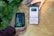 Two hands are holding objects next to some green plants. One hand holds a black and green electronic device with the brand 'Newpowa' displayed. The other hand holds a smartphone showing a product page for a 'Newpowa' 12V/24V PWM Solar Charge Controller, including product ratings and details.