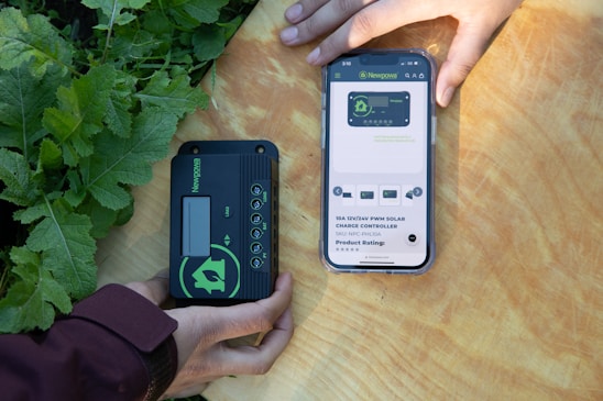 Two hands are holding objects next to some green plants. One hand holds a black and green electronic device with the brand 'Newpowa' displayed. The other hand holds a smartphone showing a product page for a 'Newpowa' 12V/24V PWM Solar Charge Controller, including product ratings and details.