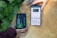 Two hands are holding objects next to some green plants. One hand holds a black and green electronic device with the brand 'Newpowa' displayed. The other hand holds a smartphone showing a product page for a 'Newpowa' 12V/24V PWM Solar Charge Controller, including product ratings and details.