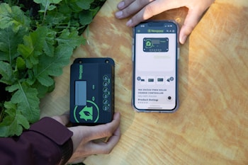 Two hands are holding objects next to some green plants. One hand holds a black and green electronic device with the brand 'Newpowa' displayed. The other hand holds a smartphone showing a product page for a 'Newpowa' 12V/24V PWM Solar Charge Controller, including product ratings and details.