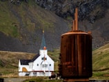 A white church with a black roof stands against a backdrop of steep, rocky hills covered in patches of green grass. In the foreground, a large, rusted industrial structure provides a stark contrast to the serene setting of the church.