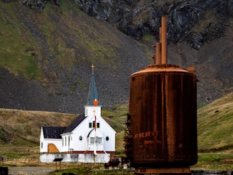 A white church with a black roof stands against a backdrop of steep, rocky hills covered in patches of green grass. In the foreground, a large, rusted industrial structure provides a stark contrast to the serene setting of the church.