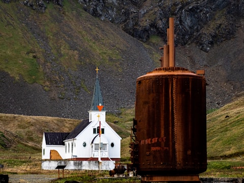 A white church with a black roof stands against a backdrop of steep, rocky hills covered in patches of green grass. In the foreground, a large, rusted industrial structure provides a stark contrast to the serene setting of the church.