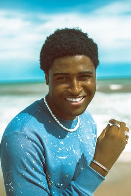 A young person smiling and standing in front of a beach. They are wearing a blue sweatshirt with paint splatters, pearl necklace, and several rings and a bracelet. The ocean and sky are visible in the background.