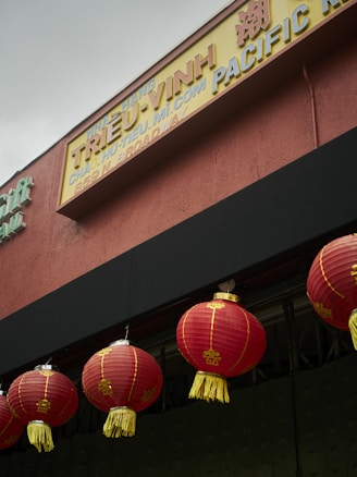 A series of red Chinese lanterns with yellow tassels hanging from the facade of a building. Above the lanterns, there is a signboard with text in both English and Vietnamese, with additional Asian script characters. The colors of the signboard are predominantly yellow and brown. The building itself has a red-brown exterior wall.