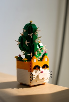 A Lego construction shaped like a cat planter with a green cactus growing out of it. The planter uses yellow, orange, and white bricks to create a cat face with pink details for the nose. The cactus is built with green bricks and has multiple protruding spines made of small beige pieces. Small flowers, also made of Lego pieces, are scattered on the cactus.