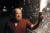 A person is holding a small firework that is actively sparking and glowing. The scene is set at night with scattered, illuminated sparks creating a bright contrast against the dark background. The individual appears captivated by the firework, and additional blurred lights can be seen in the background.