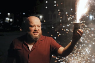 A person is holding a small firework that is actively sparking and glowing. The scene is set at night with scattered, illuminated sparks creating a bright contrast against the dark background. The individual appears captivated by the firework, and additional blurred lights can be seen in the background.