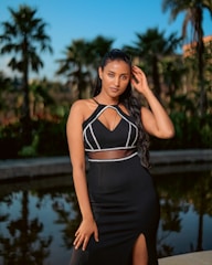 A woman poses confidently outdoors near a reflective pool, surrounded by palm trees under a clear blue sky. She is wearing an elegant black dress with cut-out details and is touching her hair with one hand, projecting an air of elegance and sophistication.