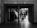 A black and white photograph reveals a dimly lit indoor passageway with tiled walls. In the middle of the frame, two people stand near a security gate or scanner, having a conversation. The area is sparse and appears to be an entrance or checkpoint.