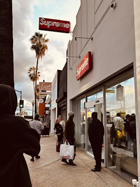People are walking on a busy city street lined with shops, including a prominent store with a red and white Supreme sign. The environment includes palm trees and a cloudy sky in the background, lending a casual urban vibe.