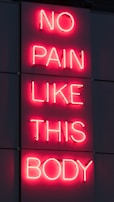 A neon sign displays the phrase 'NO PAIN LIKE THIS BODY' in bright pink letters against a dark background. The script is vertically aligned with each word on a separate line.