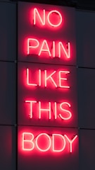 A neon sign displays the phrase 'NO PAIN LIKE THIS BODY' in bright pink letters against a dark background. The script is vertically aligned with each word on a separate line.