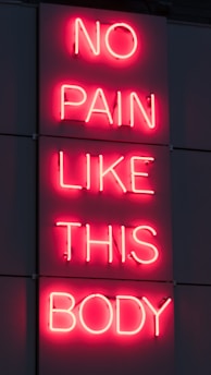 A neon sign displays the phrase 'NO PAIN LIKE THIS BODY' in bright pink letters against a dark background. The script is vertically aligned with each word on a separate line.