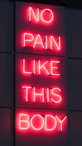 A neon sign displays the phrase 'NO PAIN LIKE THIS BODY' in bright pink letters against a dark background. The script is vertically aligned with each word on a separate line.