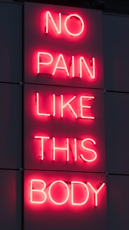 A neon sign displays the phrase 'NO PAIN LIKE THIS BODY' in bright pink letters against a dark background. The script is vertically aligned with each word on a separate line.