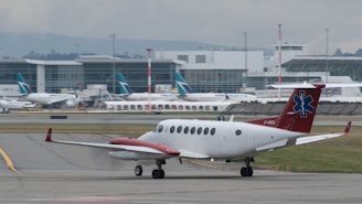 A small airplane with a red tail featuring a medical emblem is taxiing on an airport runway. In the background, a large airport terminal is visible with several other planes parked at the gates. The scene is overcast, suggesting a cloudy day.