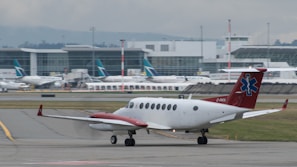 A small airplane with a red tail featuring a medical emblem is taxiing on an airport runway. In the background, a large airport terminal is visible with several other planes parked at the gates. The scene is overcast, suggesting a cloudy day.