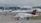 A small airplane with a red tail featuring a medical emblem is taxiing on an airport runway. In the background, a large airport terminal is visible with several other planes parked at the gates. The scene is overcast, suggesting a cloudy day.