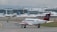 A small airplane with a red tail featuring a medical emblem is taxiing on an airport runway. In the background, a large airport terminal is visible with several other planes parked at the gates. The scene is overcast, suggesting a cloudy day.