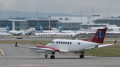A small airplane with a red tail featuring a medical emblem is taxiing on an airport runway. In the background, a large airport terminal is visible with several other planes parked at the gates. The scene is overcast, suggesting a cloudy day.