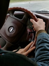 A person is gripping the steering wheel of a vehicle with both hands, with a speedometer visible in the background. The interior of the car appears to be brown with a textured steering wheel cover. The view outside the windshield includes a road and some greenery.