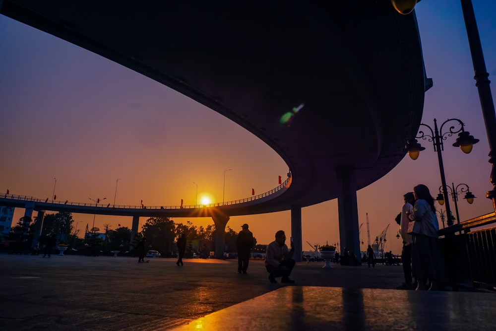 a group of people standing under a bridge at sunset
