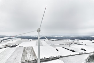 A large wind turbine stands prominently in a snow-covered landscape, with fields and scattered trees in the background. The sky is overcast, creating a serene and cold atmosphere.
