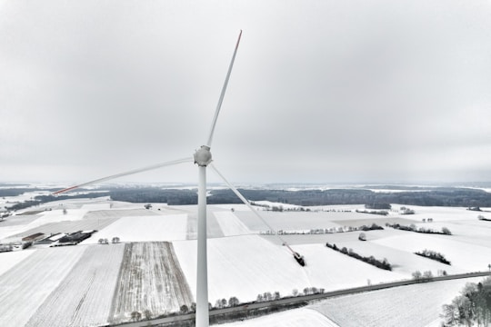 A large wind turbine stands prominently in a snow-covered landscape, with fields and scattered trees in the background. The sky is overcast, creating a serene and cold atmosphere.