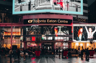 A bustling shopping center with bright digital advertisements and glass windows on the upper level. Pedestrians and vehicles move across the wet, reflective pavement of a busy urban plaza, illuminated by vibrant lights.