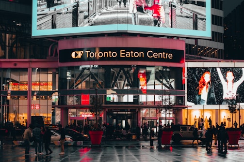 A bustling shopping center with bright digital advertisements and glass windows on the upper level. Pedestrians and vehicles move across the wet, reflective pavement of a busy urban plaza, illuminated by vibrant lights.