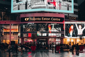 A bustling shopping center with bright digital advertisements and glass windows on the upper level. Pedestrians and vehicles move across the wet, reflective pavement of a busy urban plaza, illuminated by vibrant lights.