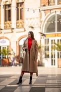 A person stands confidently in an urban setting, wearing a beige long coat over a bright red dress. They are accessorized with a tan handbag and black boots. The background features an elegant building with arched windows and decorative elements, while string lights hang above.